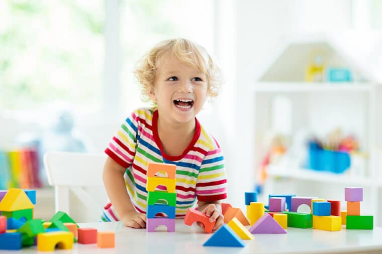 Kid playing with colorful toy blocks. Little boy building tower of block toys. Educational and creative toys and games for young children. Baby in white bedroom with rainbow bricks. Child at home.