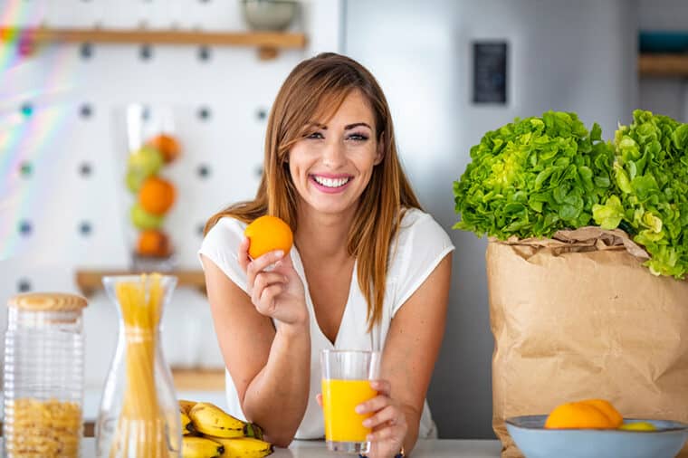 Young joyful woman drinking orange juice and standing near a kitchen table. Close up of a woman drinking juice in her kitchen. Fit smiling young woman preparing healthy fruit juice